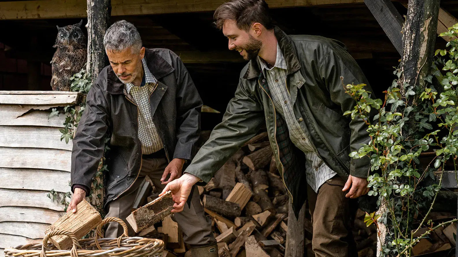 Two men stacking firewood while wearing their durable wax jackets outdoors.