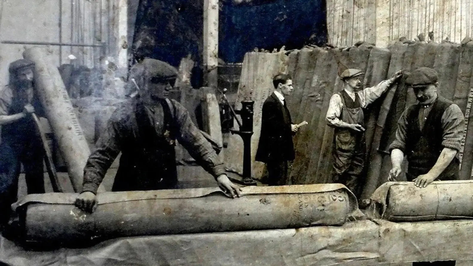 Workers handling large cylindrical objects in British Millerain waxed cotton gear.