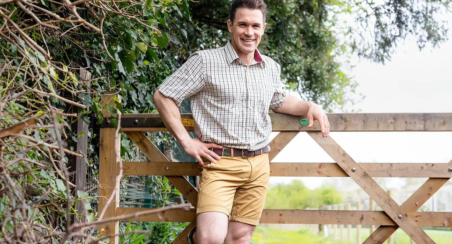 Man in a checkered short sleeve shirt and khaki shorts by a wooden gate for spring summer.