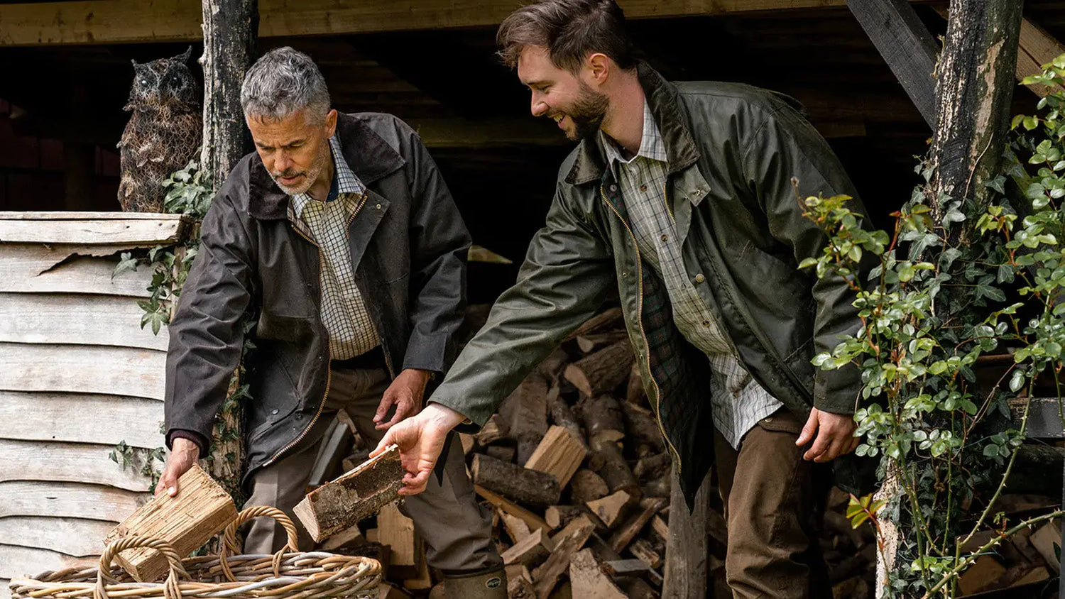 Two men stacking firewood while wearing their durable wax jackets outdoors.