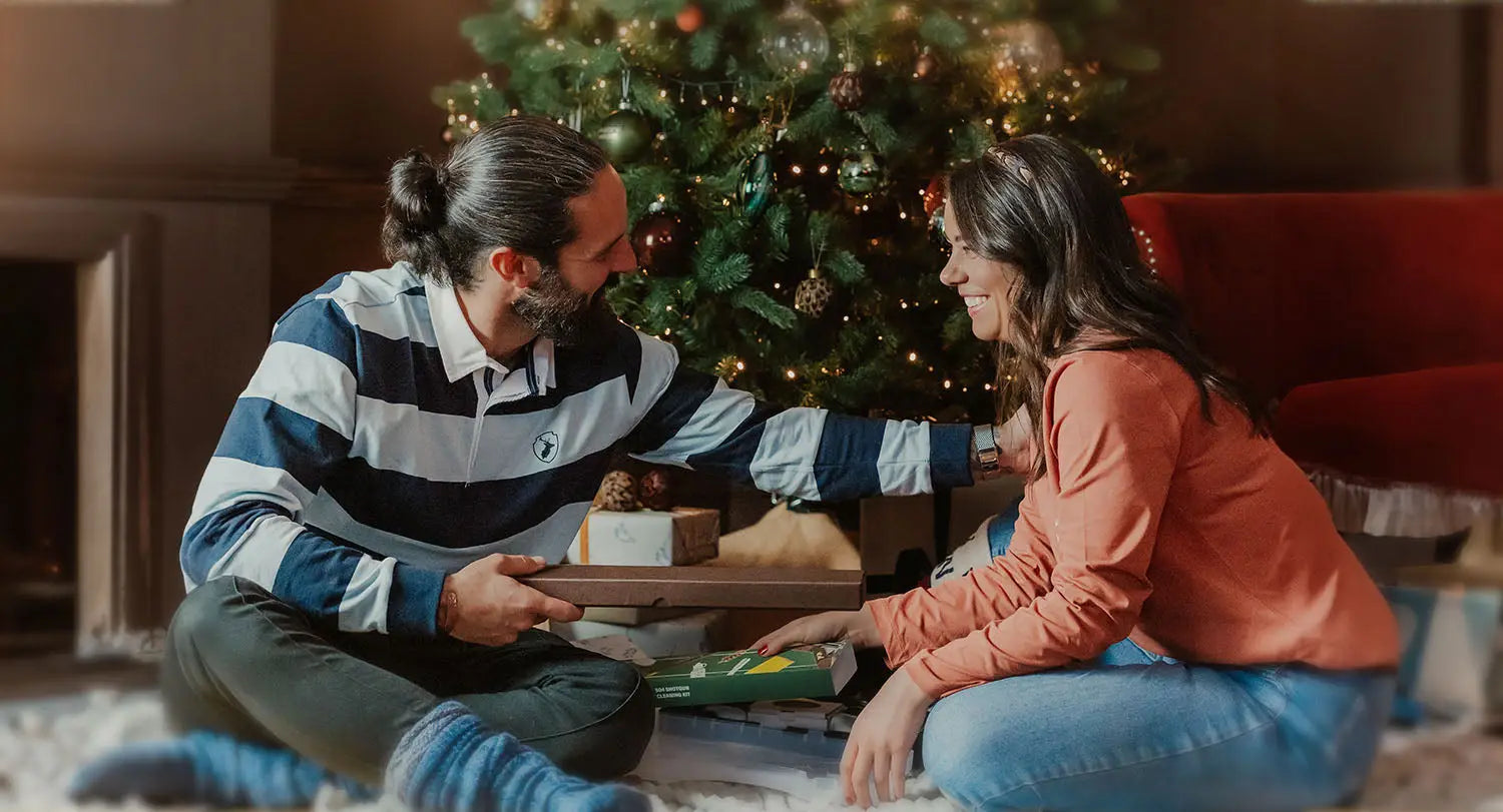 Couple exchanging gifts by a Christmas tree amidst cozy Forest Clothing offers.