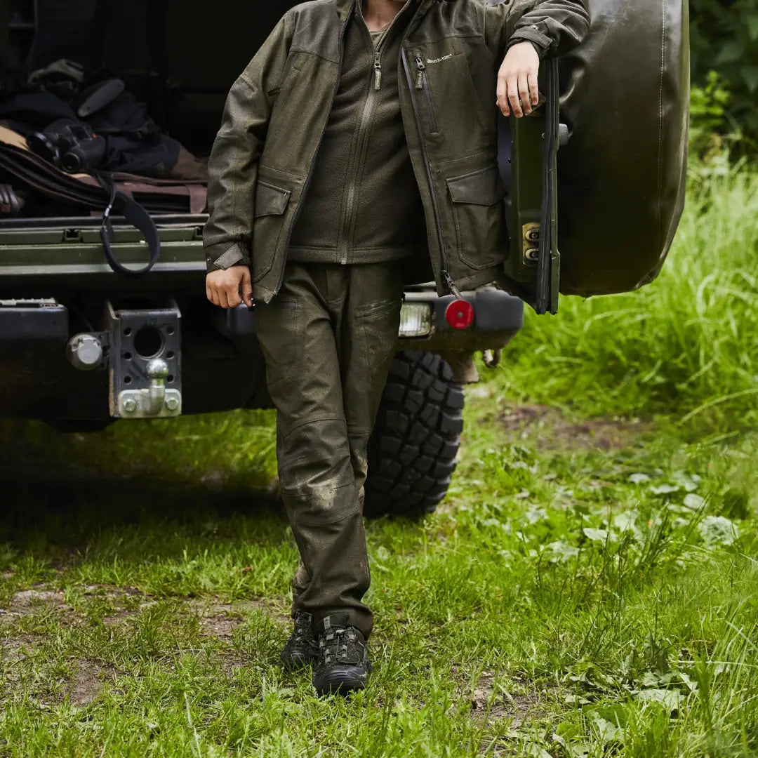 Person in olive green outdoor gear beside a vehicle showing youth chasse trousers