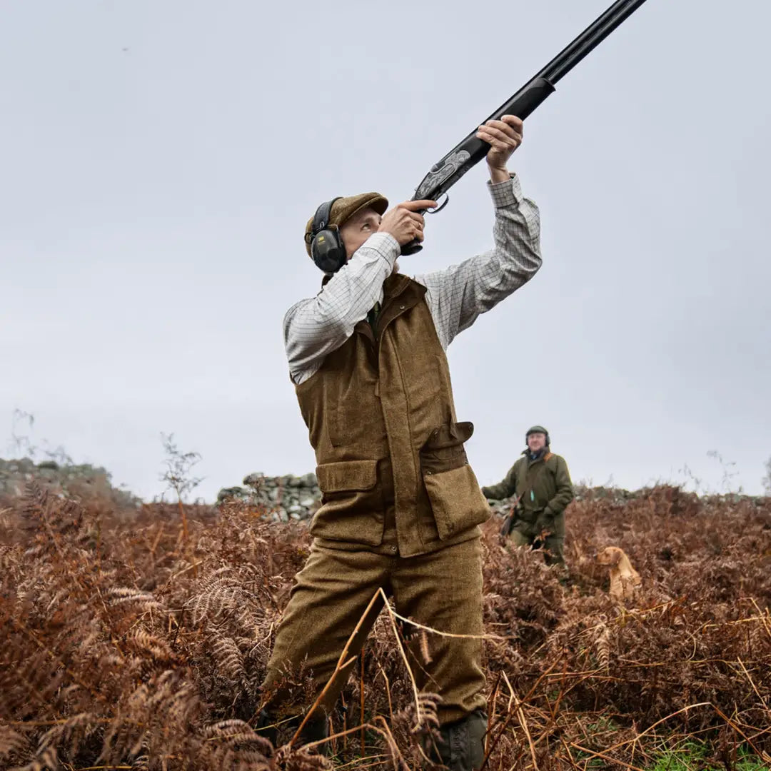 Hunter aiming a shotgun in ferns, wearing Härkila Kenmore Waistcoat with press button closure