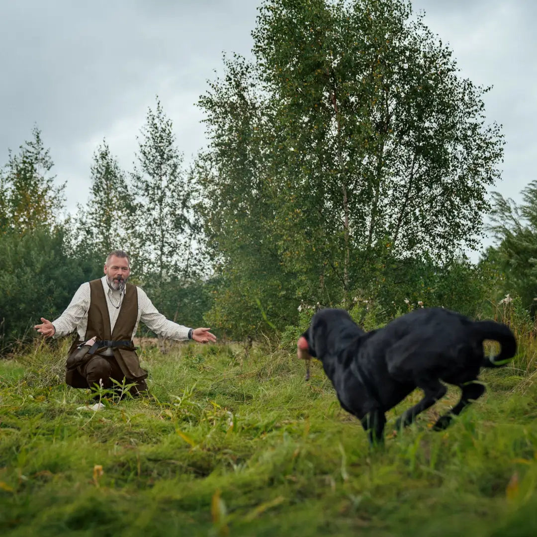 Man in vintage clothing plays with black dog in field, showcasing Retrieve Dummy Waistcoat