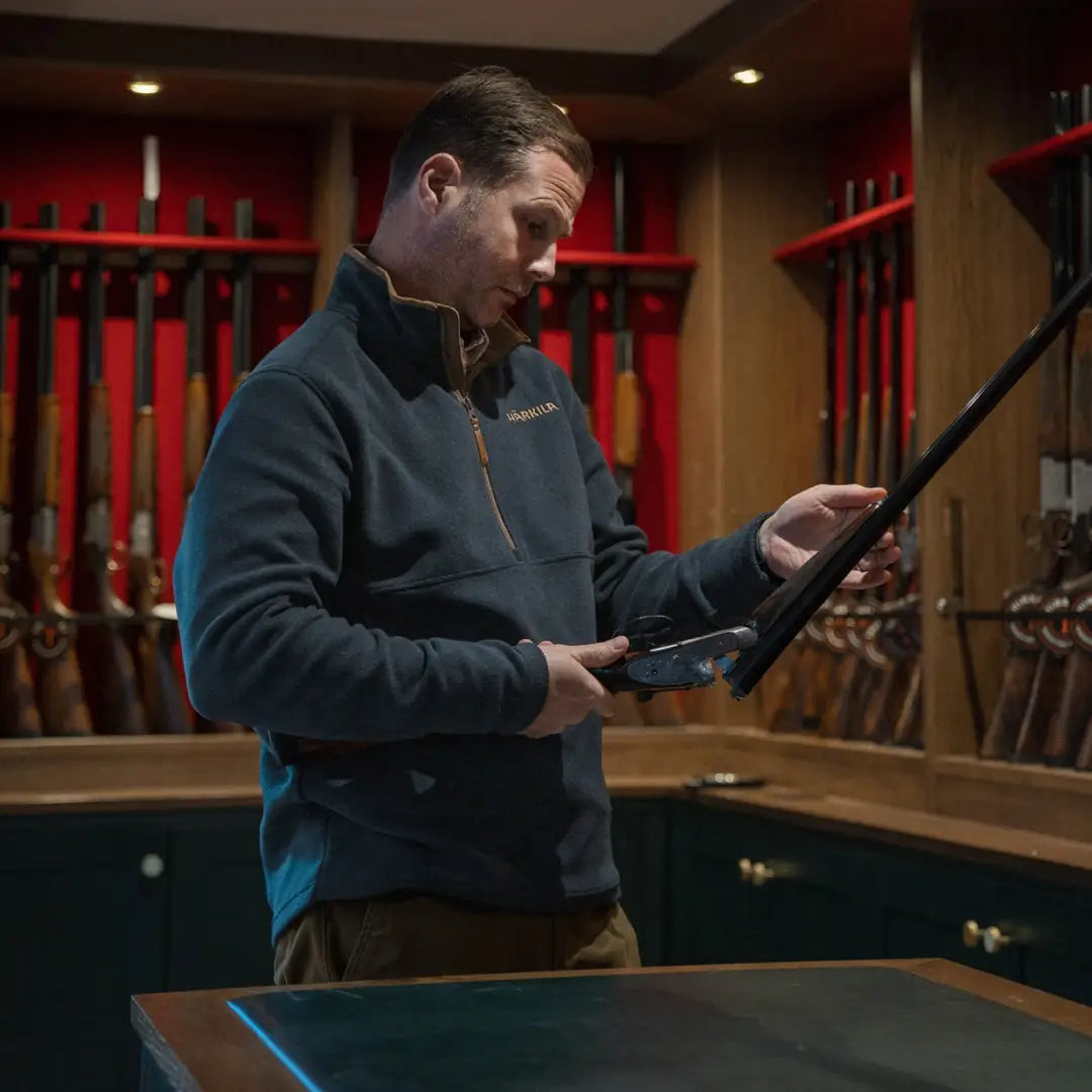 Person examining a rifle in an armory, wearing Harkila Sandhem country clothing