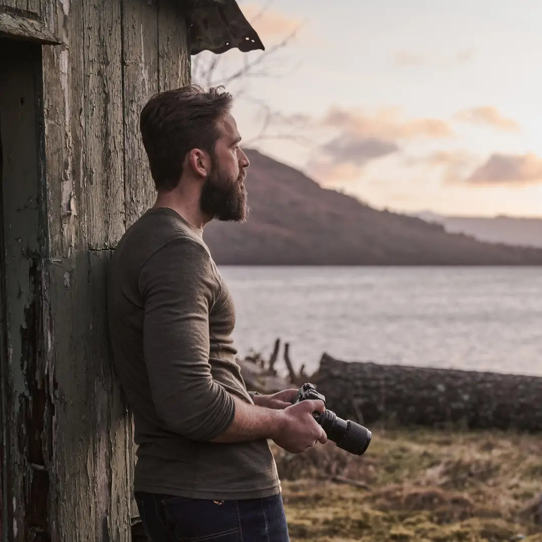 Bearded man in Merino wool crew neck top, holding a camera by a wooden structure