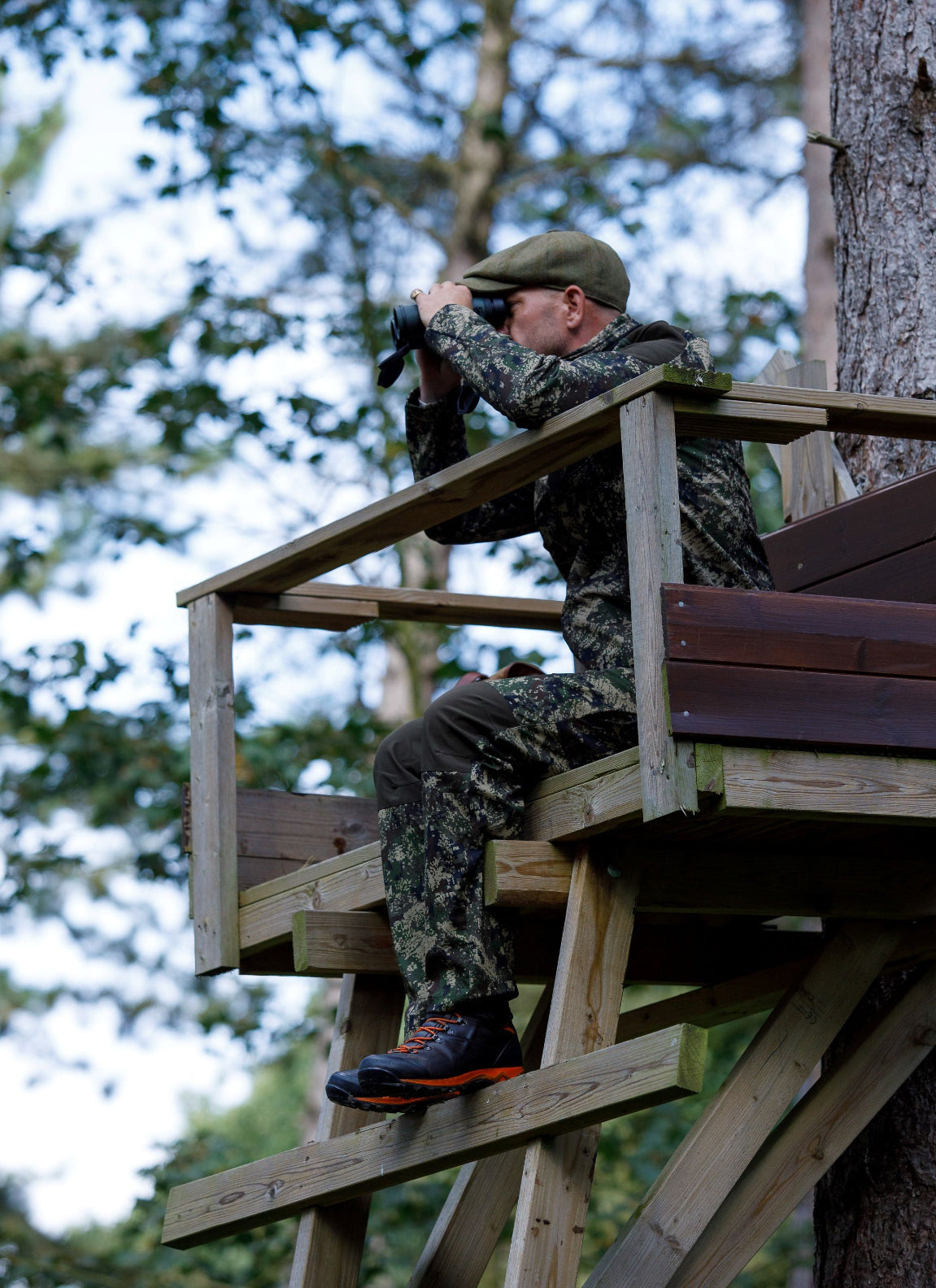 Camouflaged man in Jack Pyke Digicam Softshell Trousers using binoculars from tree stand