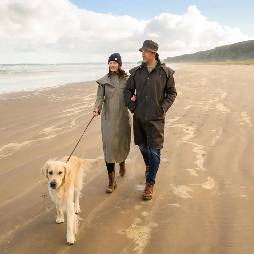 Couple in Lighthouse Stockman Full Length Waterproof Rain Coat walks golden retriever on beach