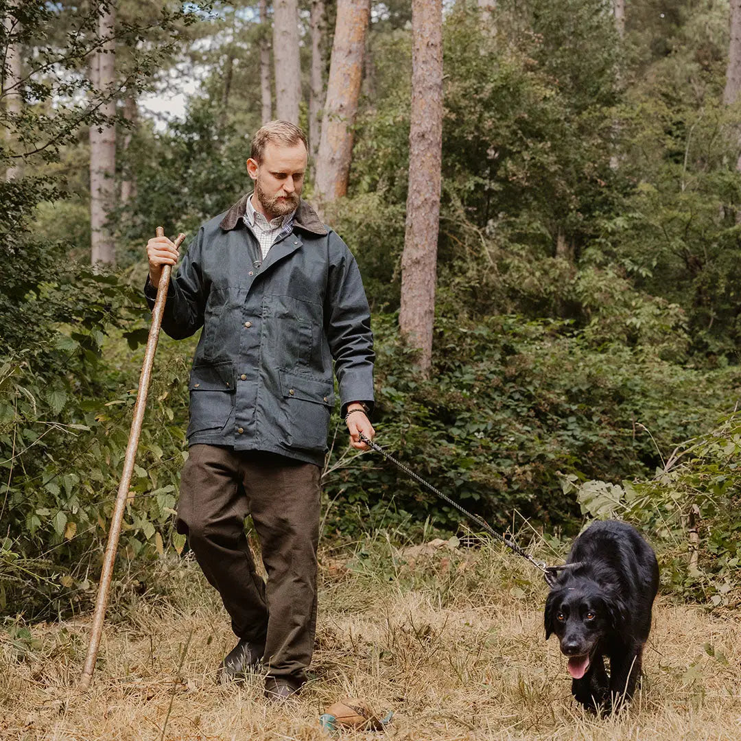 Man in a traditional wax jacket walking a black dog in a lush forest setting