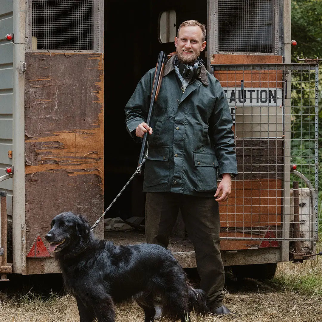 Man in a green traditional wax jacket standing with a black dog on a leash