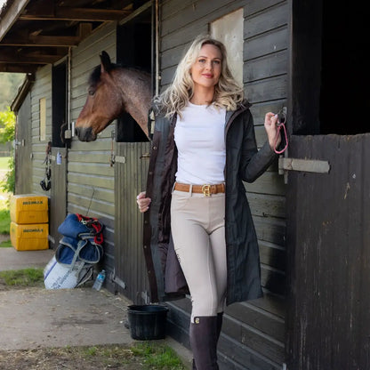 Woman in a New Forest Alice Waterproof Coat stands by a horse stable with a horse behind