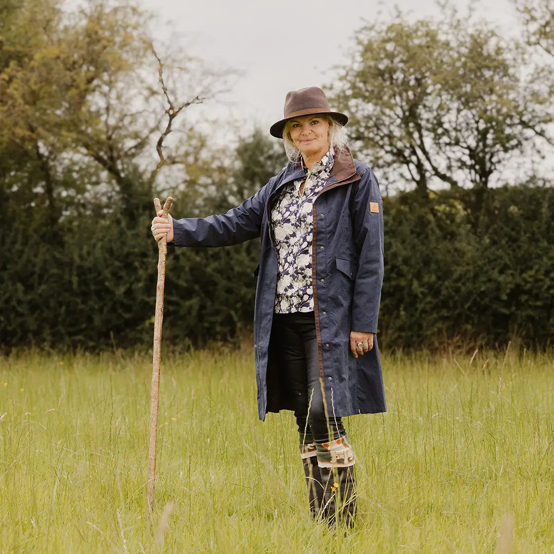 Woman in navy Forest Alice Waterproof Coat with a brown hat in a grassy field