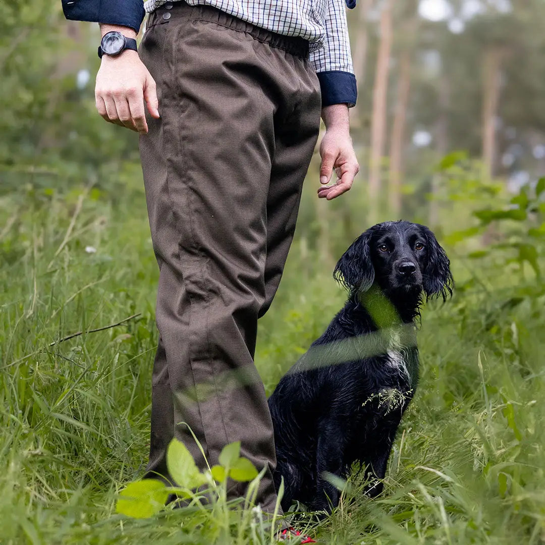 Black dog sits in tall grass by a person in Forest Hawthorn Overtrousers