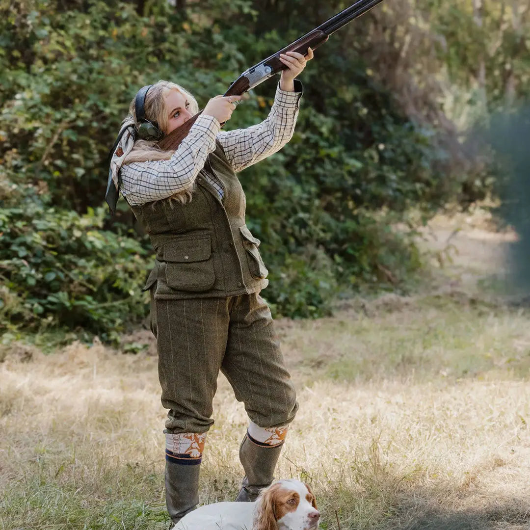 Woman in hunting gear aiming shotgun with dog, showcasing ladies tweed gilet style