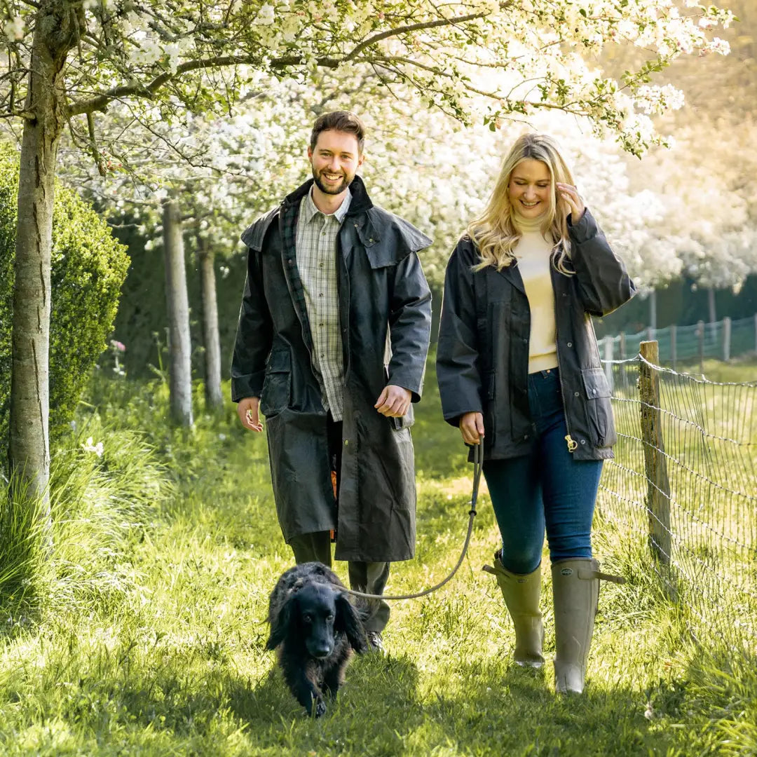 Couple in a Wax Stockman Coat walking a black dog under blooming trees