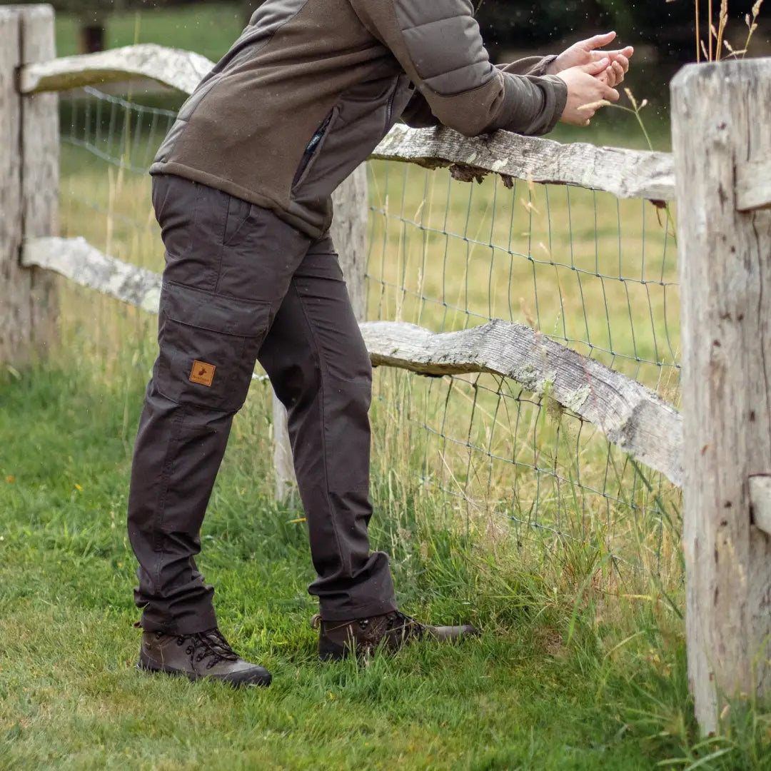 Person in New Forest Trail Trousers leaning on a rustic fence in grassy area