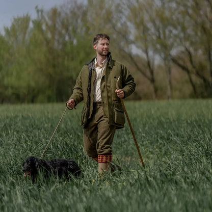 Man wearing a tweed shooting jacket walking a black dog in a grassy field