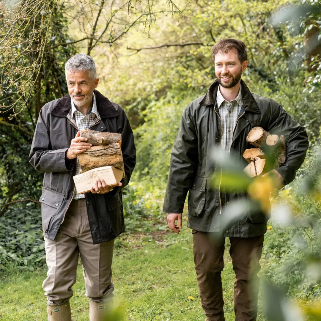 Two men in padded wax jackets walking outdoors with firewood in the forest