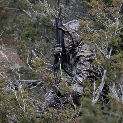 Camouflaged hunter with a rifle wearing a Percussion Hooded Sweatshirt in dense foliage