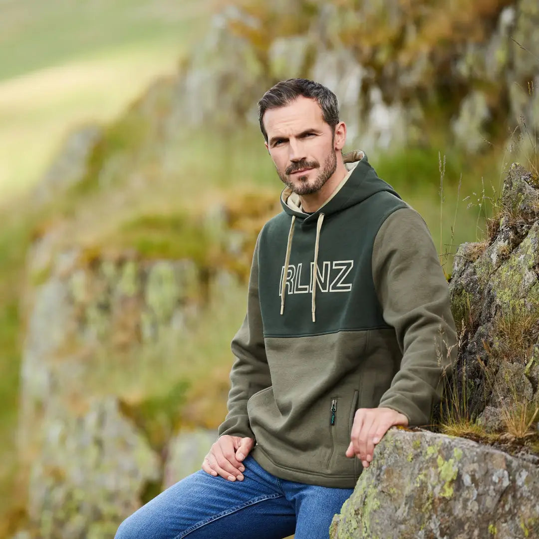 Man in a green RLNZ hoodie chilling on rocks, showcasing the South Island Hoodie style