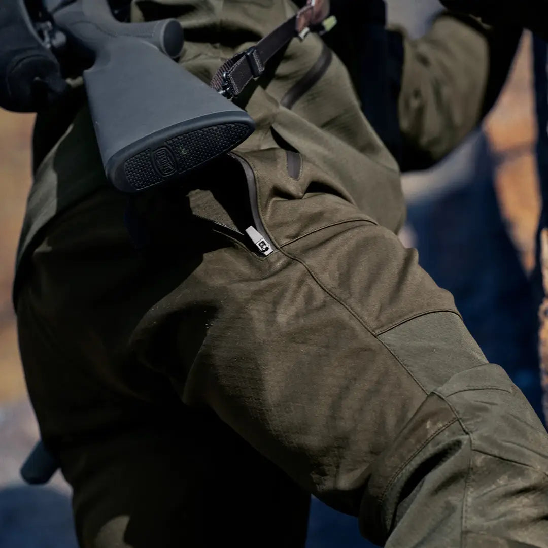 Assault rifle with black stock and olive drab body next to Seeland Hawker Shell II Trousers