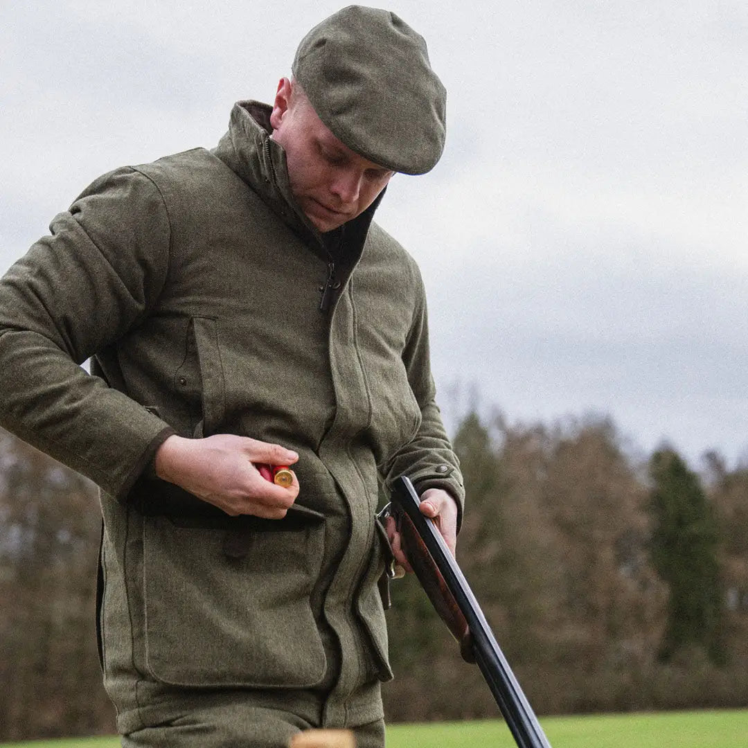 Man in olive green Seeland Hillside Jacket and flat cap, holding a shotgun