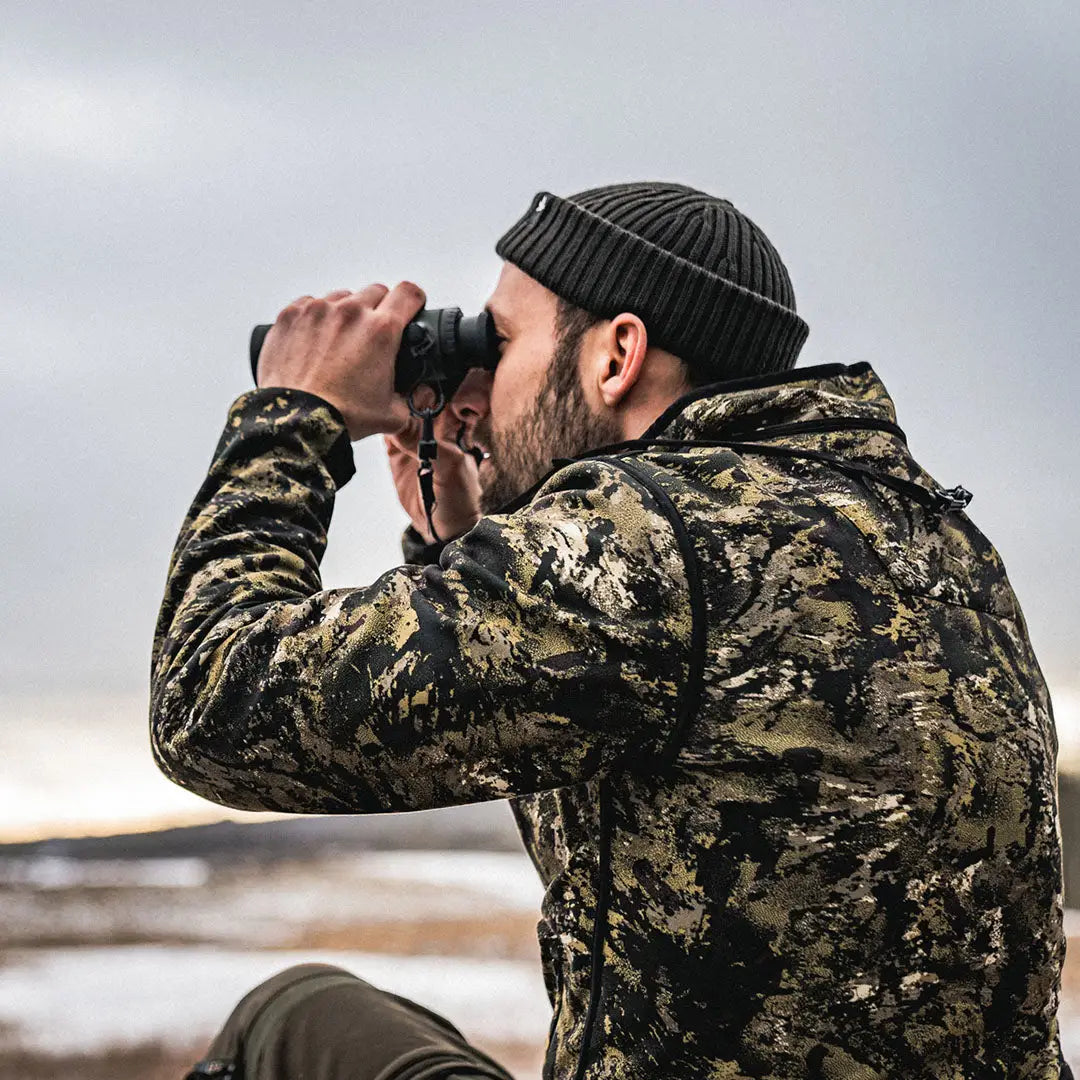 Man in camouflage with a beanie using binoculars, showcasing Seeland Rogue Reversible Fleece