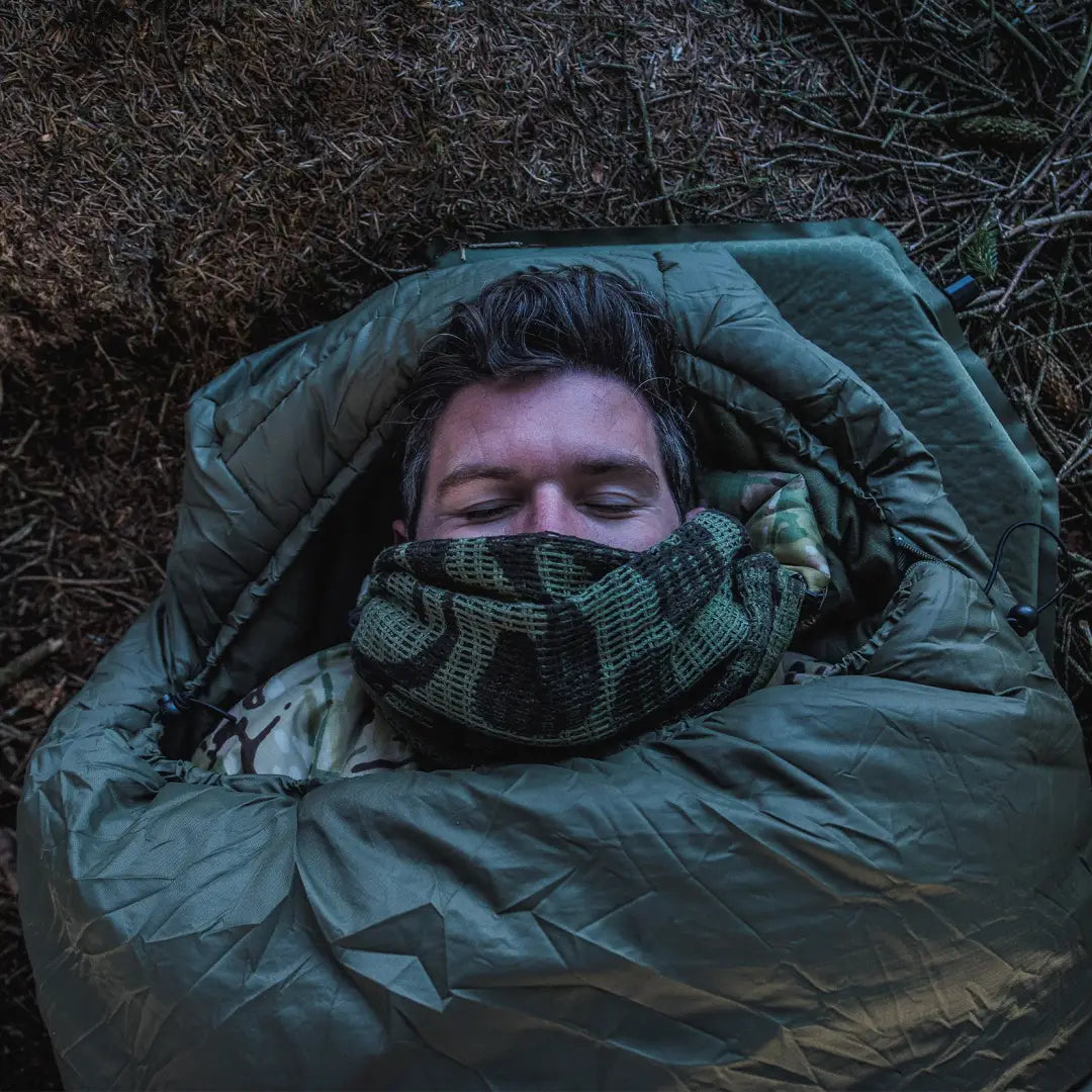 Person snoozing in a green Snugpak sleeping bag, cozy with a scarf, city lights shining