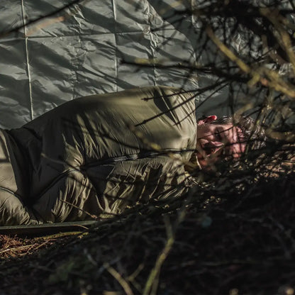 Person cozy in a green Snugpak sleeping bag amid twigs and branches