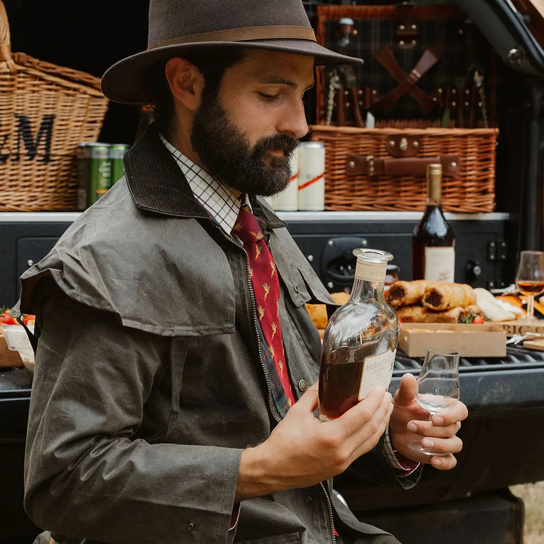 Bearded man in a hat pouring a drink while wearing a Stockman Full Length Wax Coat