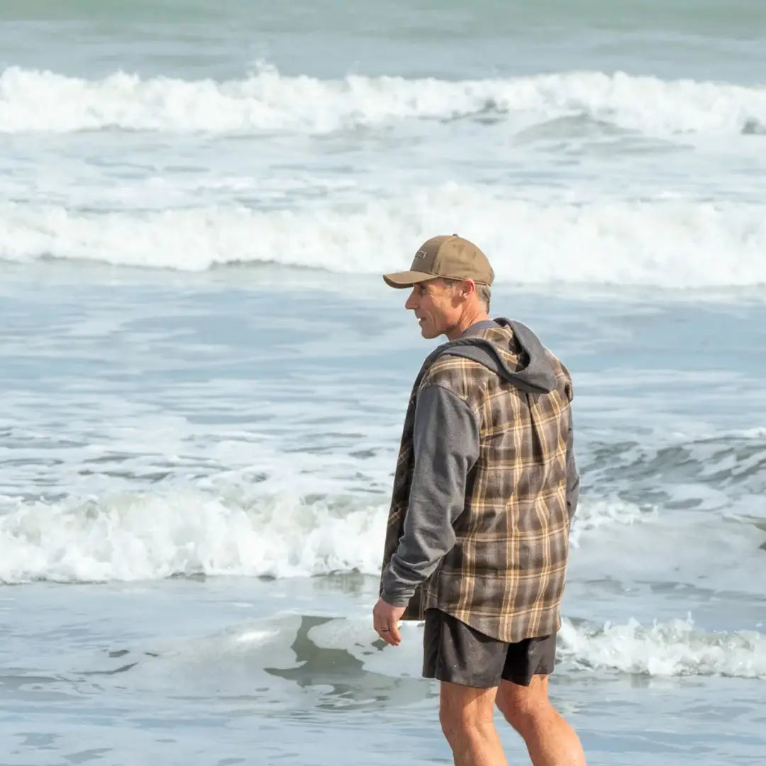 Man in plaid vest and cap at ocean, styling the Swazi Apprentice Hooded Shirt