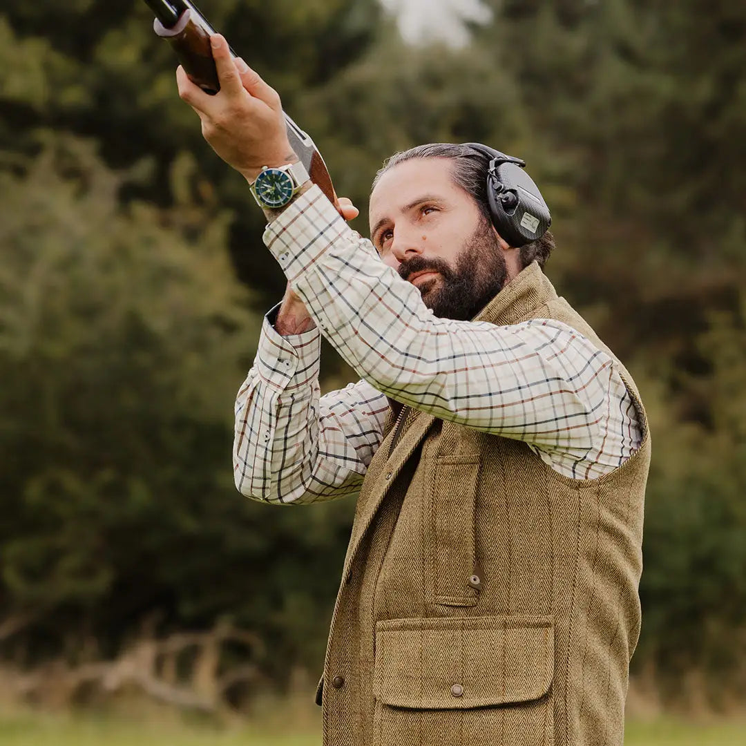 Man in tweed gilet aiming a shotgun, showcasing country clothing for hunting